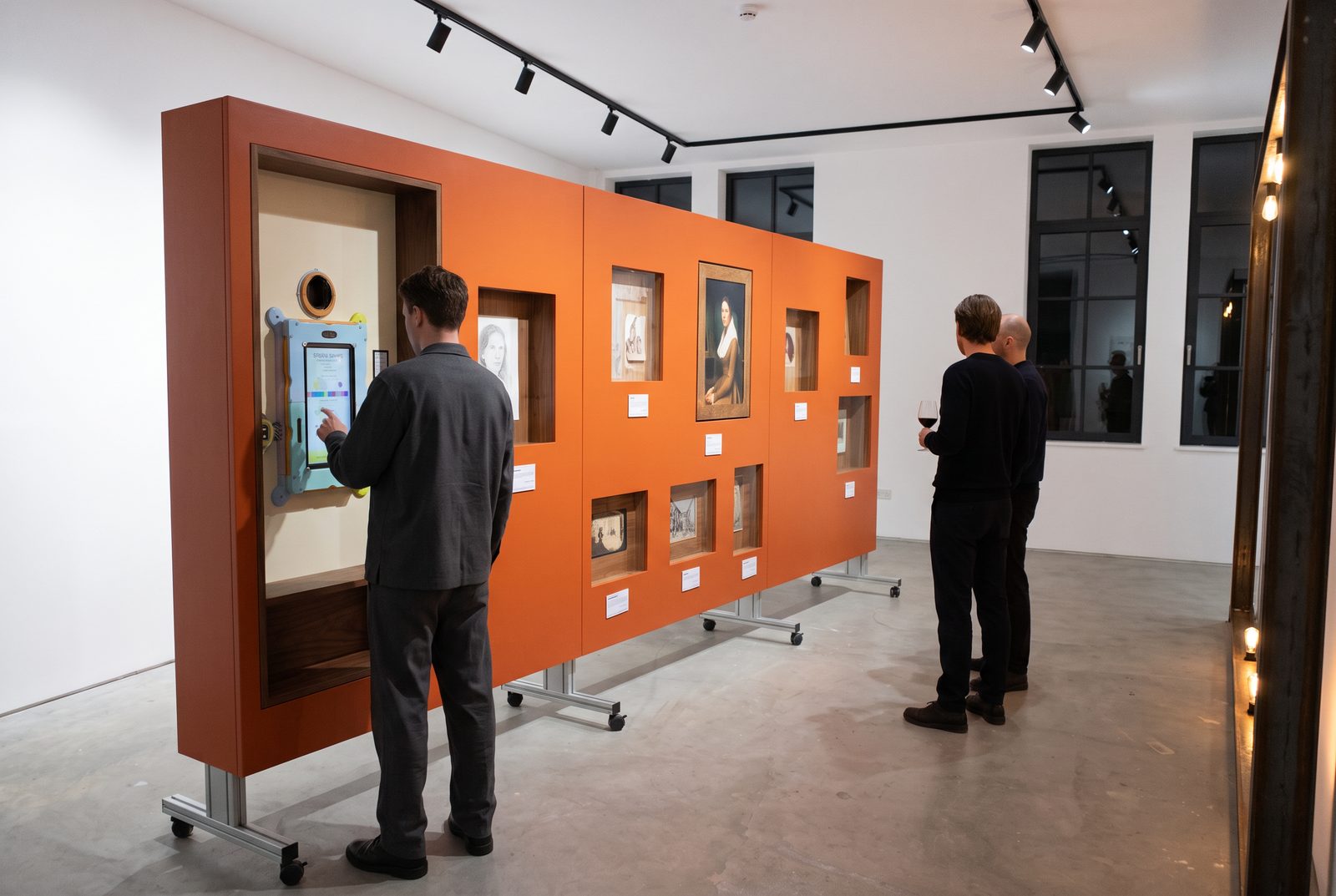 An orange exhibition wall with niches and a terminal, visitors in front of it
